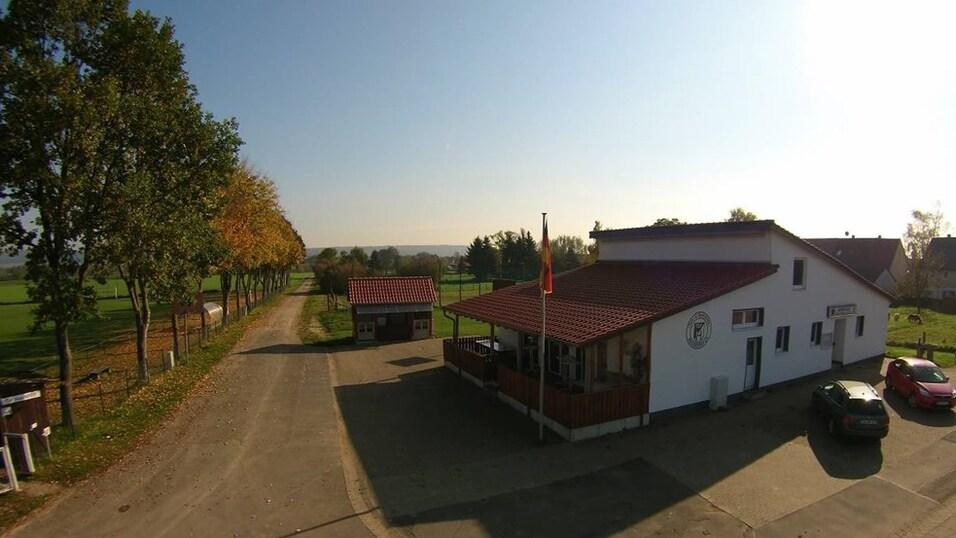 Blick auf ein wei&szlig;es Geb&auml;ude mit rotem Dach, umgeben von B&auml;umen und einem Stra&szlig;enverlauf in l&auml;ndlicher Umgebung.