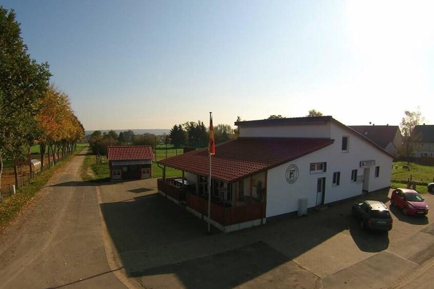 Blick auf ein wei&szlig;es Geb&auml;ude mit rotem Dach, umgeben von B&auml;umen und einem Stra&szlig;enverlauf in l&auml;ndlicher Umgebung.
