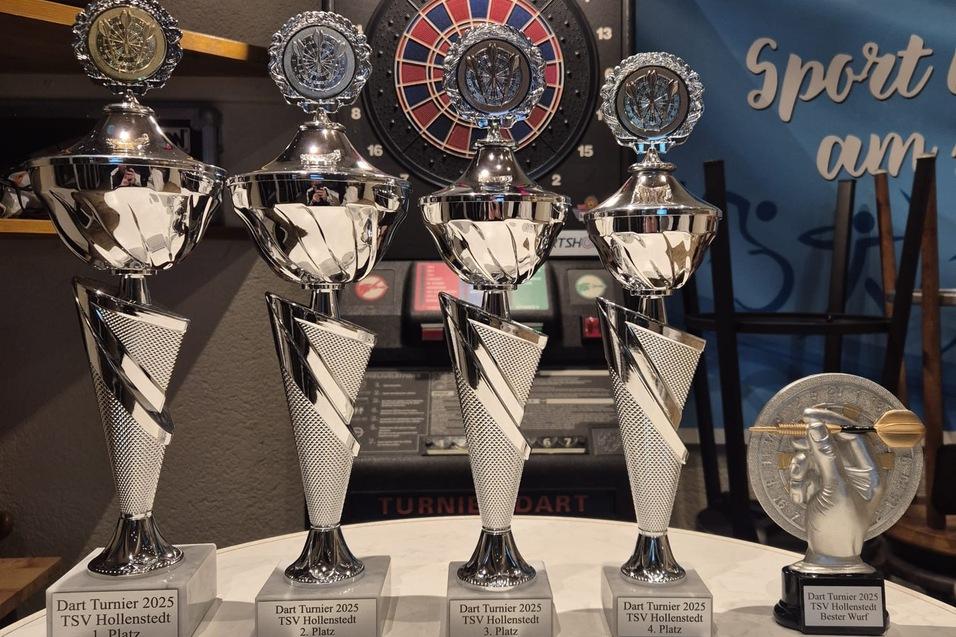 Vier silberne Pokale mit blauen Emblemen und ein kleinerer Pokal auf einem Tisch vor einer Dartscheibe und einem Sportbanner.