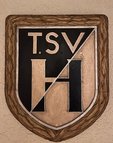 Wappen des TSV mit den Buchstaben T, S, V und H auf einem geschm&uuml;ckten Hintergrund.