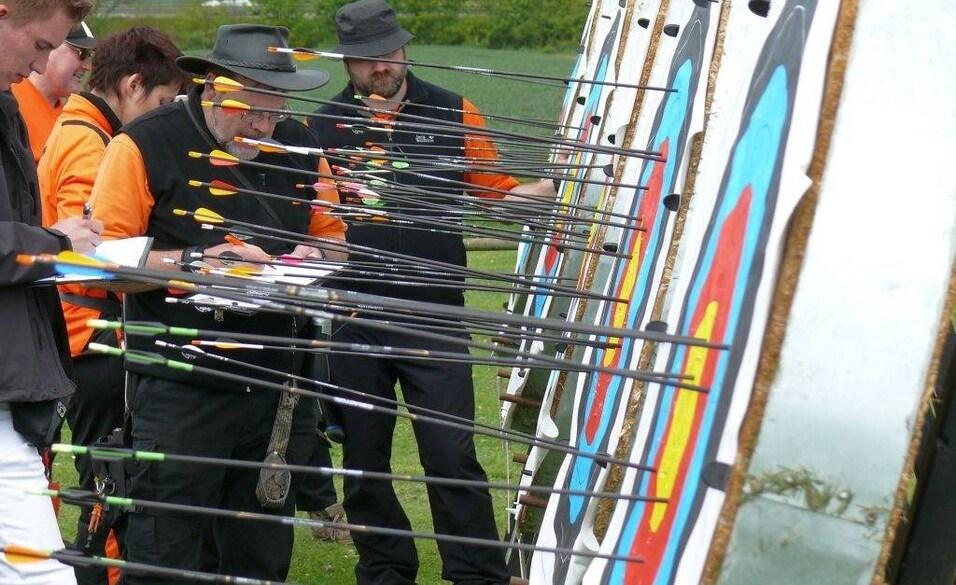 Mehrere Personen in orange-schwarzen T-Shirts notieren Ergebnisse neben Zielscheiben mit Pfeilen auf einem Feld.