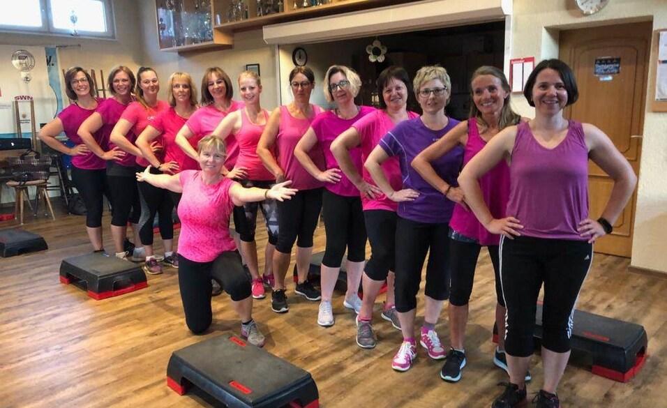 Gruppe von Frauen in Sportkleidung mit Rosat&ouml;nen posiert l&auml;chelnd vor Step-Trainingsger&auml;ten in einem Fitnessraum.