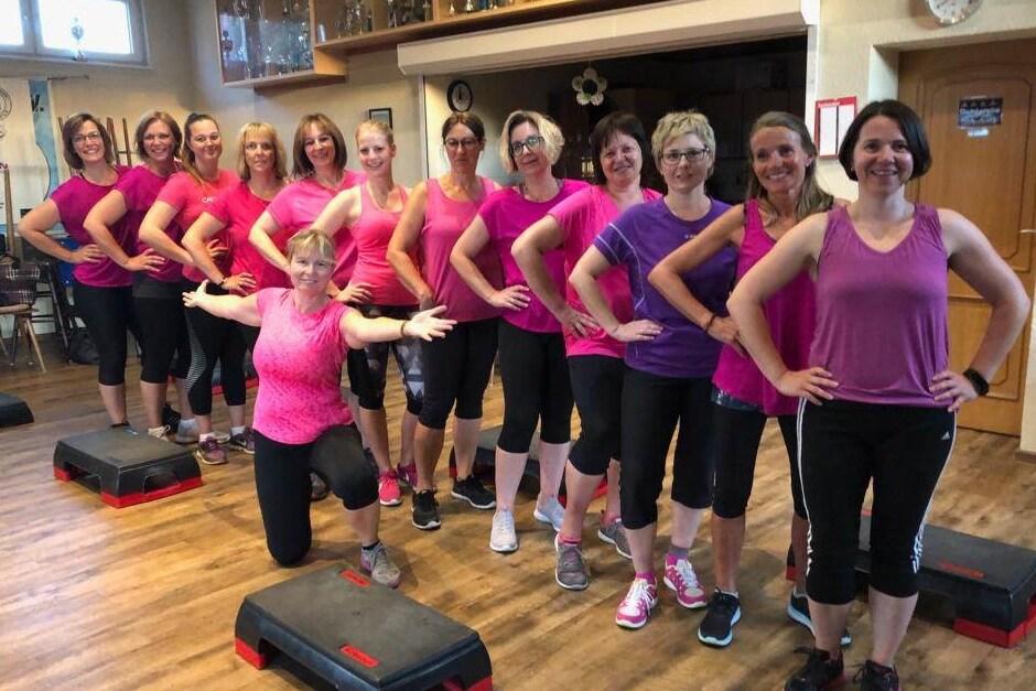 Gruppe von Frauen in Sportkleidung mit Rosat&ouml;nen posiert l&auml;chelnd vor Step-Trainingsger&auml;ten in einem Fitnessraum.