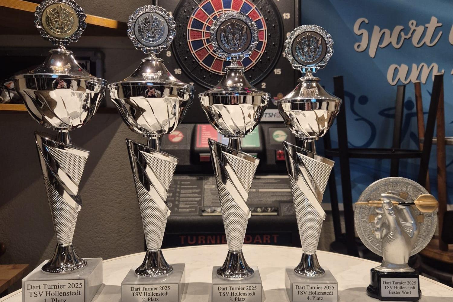 Vier silberne Pokale mit blauen Emblemen und ein kleinerer Pokal auf einem Tisch vor einer Dartscheibe und einem Sportbanner.