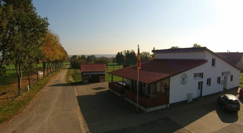 Landschaftsansicht eines Geb&auml;udes mit rotem Dach und deutscher Flagge an einer Landstra&szlig;e, umgeben von B&auml;umen.