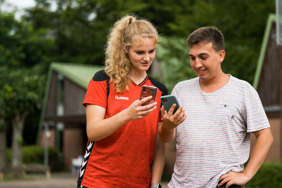 Zwei junge Leute schauen auf ihre Smartphones, stehen lächelnd im Freien vor einem Gebäude.