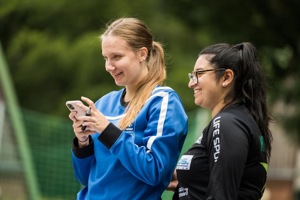Zwei Frauen lächeln, während eine auf ihrem Smartphone tippt, umgeben von grüner Natur und Sportgeräten im Hintergrund.