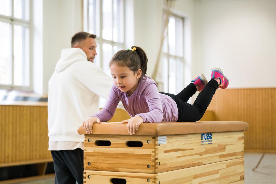 Ein Mädchen liegt auf einem Turnkasten, während ein Trainer im Hintergrund beobachtet. Die Umgebung ist ein Sportraum.